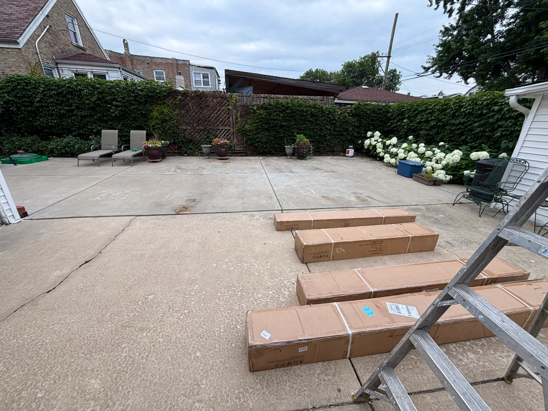 Before assembly: the open patio with packaged gazebo components and a ladder ready at the work area.