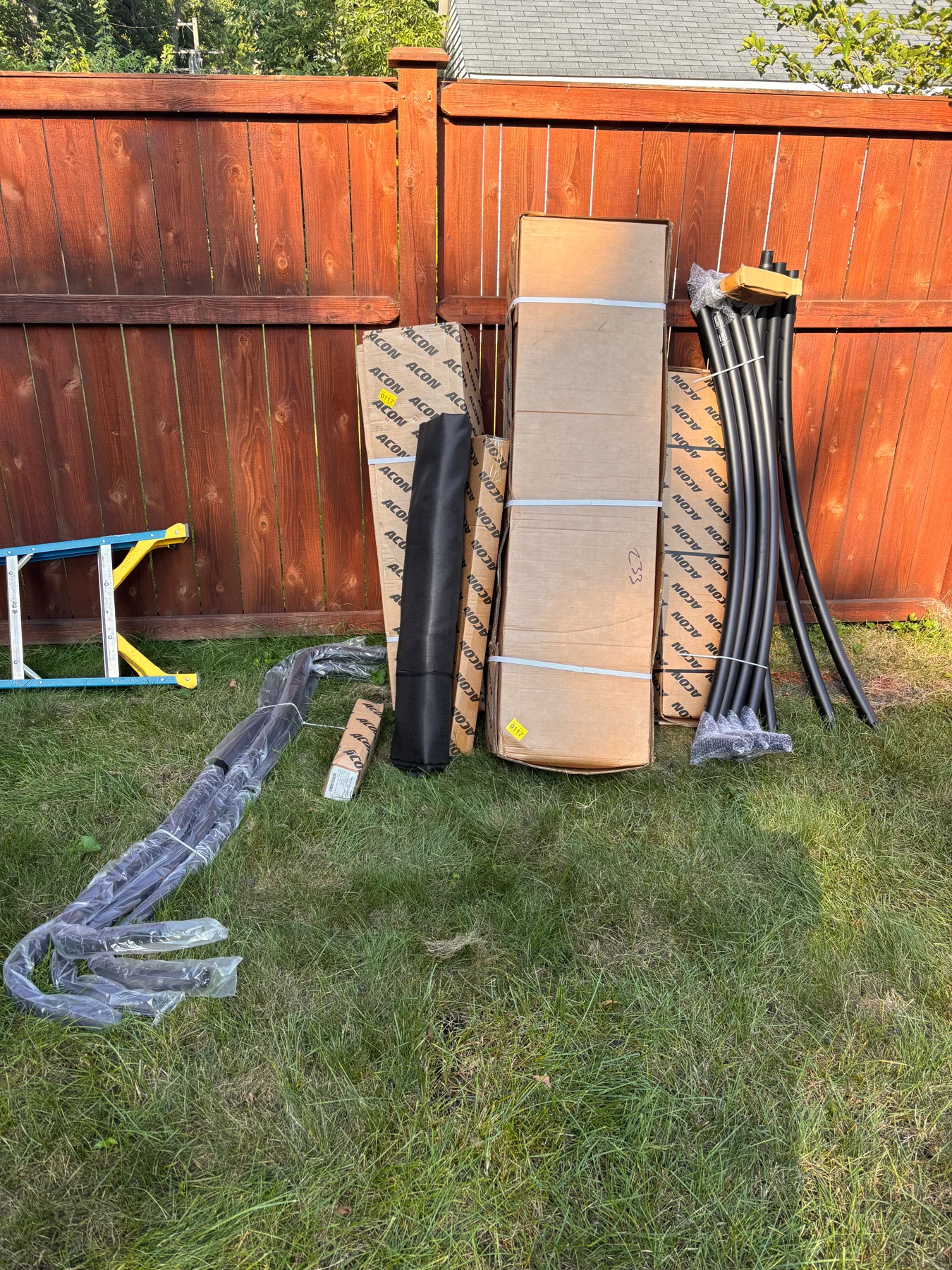 Before assembly: boxes, poles, and other trampoline components arranged beside the garden fence.